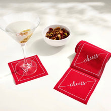 Load image into Gallery viewer, Roll of red cotton cocktail napkins with a white border and "cheers" in white script displayed on a white surface beside a martini resting on a red "cheers" cocktail napkin and a white snack bowl filled with snacks.
