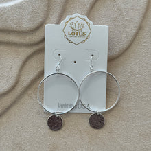 Load image into Gallery viewer, Pair of sterling silver hoop earrings featuring a hand-hammered round disc charm on each, displayed on a tan textured surface.