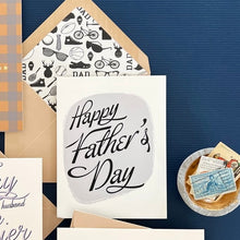 Load image into Gallery viewer, Happy Father's Day Card, cream greeting card with black script text, on gray center panel, beige envelope with black and white specialized dad graphics liner, shown with coordinating paper pieces, displayed on blue surface. 