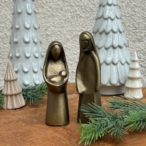 Nativity set crafted from cast aluminum with an antique gold finish. Behind the set are two ,tall, white -colored, Christmas trees ; and two, small, cream-colored, Christmas trees. On a wooden surface garnished with pine needle cuttings and against a white textured background. 