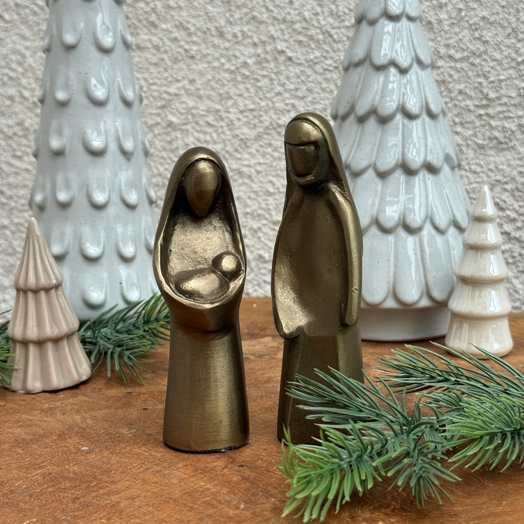 Nativity set crafted from cast aluminum with an antique gold finish. Behind the set are two ,tall, white -colored, Christmas trees ; and two, small, cream-colored, Christmas trees. On a wooden surface garnished with pine needle cuttings and against a white textured background. 