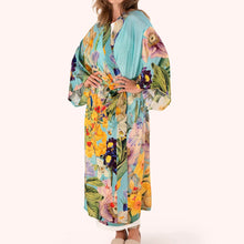 Load image into Gallery viewer, Spring Flora Kimono Gown worn by model, lightweight fabric with large floral pattern in yellow, green, and purple tones on a turquoise-blue background, tie belt, wide sleeves, full-length silhouette, against a white background. 