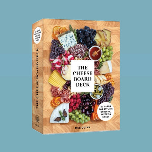 Box cover of The Cheese Board Deck by Meg Quinn featuring an overhead view of a colorful charcuterie board with assorted cheeses, meats, fruits, honey, and dried oranges arranged on a wooden surface, with a white square at the center displaying the title in black lettering. Book against a light blue background. 