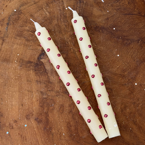 Two white unlit candles with metallic red polka dots, lying side by side, on a brown wooden surface.
