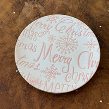 Load image into Gallery viewer, A white round stoneware plate with  &quot;Merry Christmas&quot; in red. &quot;Merry Christmas&quot; is repeated and various lettering and sequencing. Plate is on a brown wooden surface. 