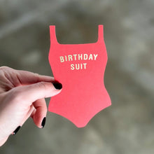 Load image into Gallery viewer, Birthday Suit Flat Greeting Card, swimsuit-shaped greeting card in bright coral with gold Birthday Suit text, held in hand against a blurred background.