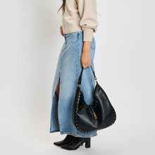 Load image into Gallery viewer, Model carrying a black pebbled vegan-leather hobo bag with gold stud embellishments and gold hardware, styled with a cream-colored sweater, light wash denim skirt, and black heeled boots against a white background. 