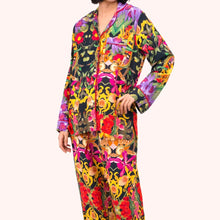Load image into Gallery viewer, Botanical Floral Pyjamas, model wearing a long-sleeve viscose button-front top and matching trousers featuring a vibrant multi-colored floral rainforest pattern on a black background, finished with hot pink piping and a chest pocket against against a white background. 