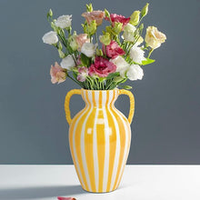 Load image into Gallery viewer, Large Yellow Striped Urn Vase, glossy yellow and white ceramic urn vase with a cinched waist, vertical stripes, flared neck, and yellow rope-style side handles, displayed with pink and white flowers on a white surface against a gray background. 