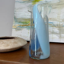Load image into Gallery viewer, Tall clear glass pitcher with blue and light blue elongated accents and pour spout, displayed on a wooden surface in front of abstract painting.