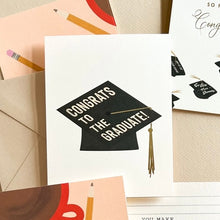 Load image into Gallery viewer, Congrats To The Graduate Card, white graduation card with black graduation cap design, gold foil tassel accents, Congrats To The Graduate white text, and coordinating graduation cards in background against neutral surface. 