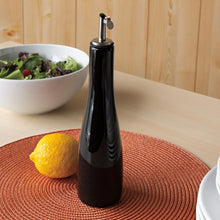 Load image into Gallery viewer, Black ceramic cruet bottle with stainless steel pour spout, displayed on an round orange placemat with lemon and white bowl of salad in the background. 