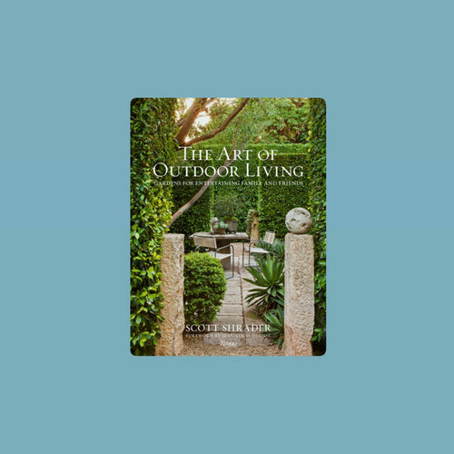 Book cover of The Art of Outdoor Living by Scott Shrader featuring a lush garden pathway framed by stone pillars and greenery leading to an outdoor seating area surrounded by manicured hedges. Book title in white lettering. Book against a light blue background.
