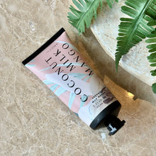 Load image into Gallery viewer, Coconut Milk Mango Demi Hand Cream, pale pink and pale aqua botanical accents with a closed black cap and crimped end displayed beside green fern fronds on a beige marble surface. 