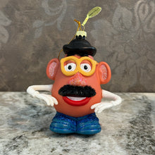 Load image into Gallery viewer, Potato Head Ornament, lifestyle image of an orange-brown potato-shaped character with yellow glasses, a black hat, a textured black mustache, glittery pink nose and ears, white arms and gloves, red smiling lips, and blue glitter-accented shoes, displayed on a gray stone surface against a gray patterned background.