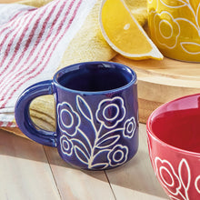 Load image into Gallery viewer, Zoey stoneware espresso cup in deep blue with white floral line design and matching blue handle, displayed on a wooden surface with a coordinating red and yellow cups, citrus slices, and red striped kitchen towel in the background.