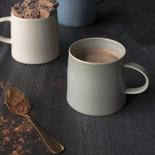 Load image into Gallery viewer, Aquarius Sage stoneware mug with reactive glaze filled with hot chocolate, on a dark wood surface with gold spoon and cocoa powder. 