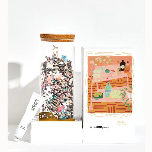 White opened box with a clear jar with a cork top filled with jigsaw puzzle pieces. On the box top there's a picture of a pink bar cart with bottles and flowers on it. Propped to the side is a white tube of jigsaw glue. White background. 