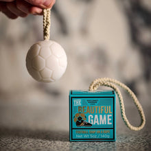 Load image into Gallery viewer, Football Ball Soap On A Rope, hand holding white soccer ball-shaped soap on a cream rope beside square turquoise and black packaging box with Beautiful Game white and gold lettering and soccer ball and cleat illustration, with cream rope extending from the box onto a textured surface. 