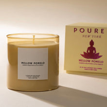 Load image into Gallery viewer, Pour NY Mellow Pomelo candle with cream wax in translucent golden glass vessel with cream packaging box featuring metallic pink lotus figure on a white surface. 