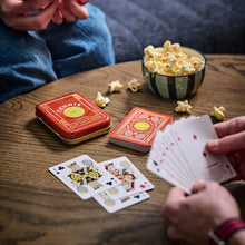 Load image into Gallery viewer, Tennis Themed Playing Cards, red tennis-themed playing card tin, stacked red card deck, and hand holding fanned playing cards displayed on a wooden table beside a striped bowl of popcorn and three face-up playing cards. 