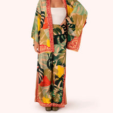 Load image into Gallery viewer, Exotic Leaves Wide Leg Trousers worn by model, lightweight fabric with large tropical leaf pattern in green, yellow, and coral tones, red patterned trim, wide leg silhouette, paired with matching kimono, against a white background. 