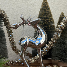 Load image into Gallery viewer, A silver metallic deer ornament with a silver looped string around the antlers. Three green decorative Christmas trees and a strand of silver glitter garland behind the deer. Against a white textured surface. 
