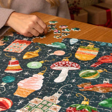 Load image into Gallery viewer, Partially assembled vintage-styled Christmas ornament jigsaw puzzle on a wooden table with a hand placing pieces near the top of the puzzle. 