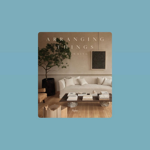 Book cover of Arranging Things by Colin King featuring a minimalist living room with a white sofa, wooden coffee table stacked with white books, and a tall leafy potted tree against a neutral beige wall. Book title and author in white lettering. Book against a light blue background.  