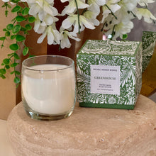 Load image into Gallery viewer, Michel Design Works Greenhouse candle with white wax and double wick in clear glass vessel beside green box with white botanical pattern, displayed on a round stone surface with white floral background. 