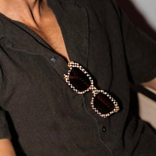 Load image into Gallery viewer, Demi Toasted Moxie Checkered Sunglasses, square textured acetate sunglasses with brown, cream, and amber checkered frames and dark brown lenses tucked into the neckline of a dark charcoal button-up shirt in a lifestyle setting. 