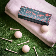Load image into Gallery viewer, Golf Ball Soaps, rectangular dark green product box with coral pink vintage-style lettering and golf illustrations, displayed with three cream -colored golf ball soaps and white golf tees on artificial green grass beside folded cream towel. 