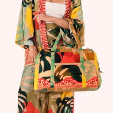 Load image into Gallery viewer, Luxe Exotic Leaves Weekender Bag worn by model with matching kimono and trousers, structured duffel with large bold tropical leaf pattern in green, coral, and mustard tones, double handles and detachable shoulder strap, front zip pocket, against a white background.