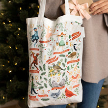 Load image into Gallery viewer, Cotton canvas tote bag featuring the 12 Days of Christmas illustrations, shown being held in front of a softly blurred lit Christmas tree. 