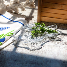 Load image into Gallery viewer, Swimming Fish Dish, clear fish-shaped glass dish with raised scale detailing, displayed with fresh green herbs on a light stone surface beside a blue and green textile, white coral accent, and a wooden decorative element. 