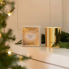 Load image into Gallery viewer, Metallic gold scented candle and its decorative white and gold box displayed on a white surface with evergreen sprigs, with a blurred lit Christmas tree in the foreground. 