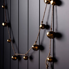 Load image into Gallery viewer, Jingle Bell Garland, small and large round brass jingle bells tied along a brown twine and loosely draped across a dark paneled wall. 