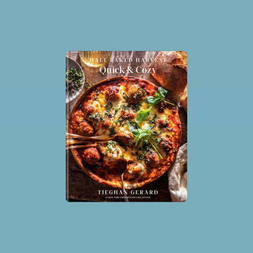 Cookbook cover of Half Baked Harvest: Quick & Cozy by Tieghan Gerard, featuring a golden-brown baked dish topped with melted cheese, herbs, and basil leaves on a tabletop. Title in white lettering. Book against a light blue background.  