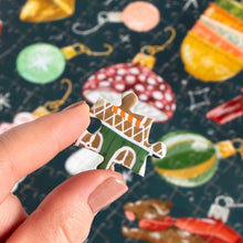 Load image into Gallery viewer, Close-up of a hand holding a single illustrated puzzle piece above the completed vintage-style Christmas ornament jigsaw puzzle in soft focus. 