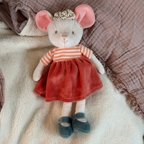 Plush mouse with soft beige fur, pink inner ears, and black stitched eyes, wearing a white bow headband with teal polka dots, orange-and-white striped shirt, orange velvet skirt, and teal shoes. Mouse is lying on a soft white blanket with a mauve fringed textile in the background. 