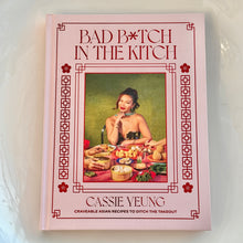 Load image into Gallery viewer, Bad B*tch In The Kitchen by Cassie Yeung, pale pink hardcover cookbook with portrait cover illustration, red geometric border details, and floral corner motifs against white background. 