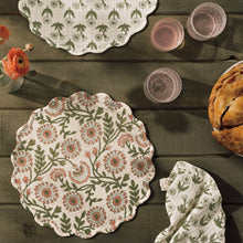 Load image into Gallery viewer, Thicket Placemat, cotton placemat with botanical floral print in peach and olive on a cream background with scalloped border, displayed on a wooden surface with coordinating placemats, glasses, and food. 