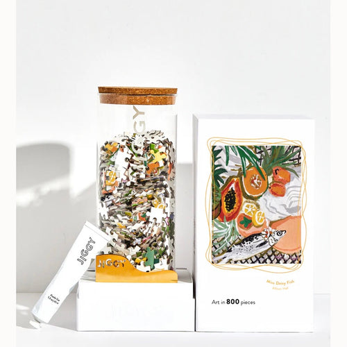 White opened box with a clear jar with a cork top filled with jigsaw puzzle pieces. On the box top there's a picture of papayas and fish on a nature background. Propped to the side is a white tube of jigsaw glue. White background. 