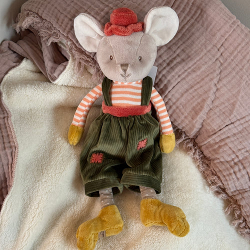Plush mouse with soft beige fur, floppy ears, and black stitched eyes, wearing a coral velvet hat, coral-and-white striped shirt, green corduroy overalls with a coral patch on each pant leg and a matching coral waistband. The mouse has mustard-colored hands and feet and is lying on a soft white blanket with a mauve fringed textile in the background. 