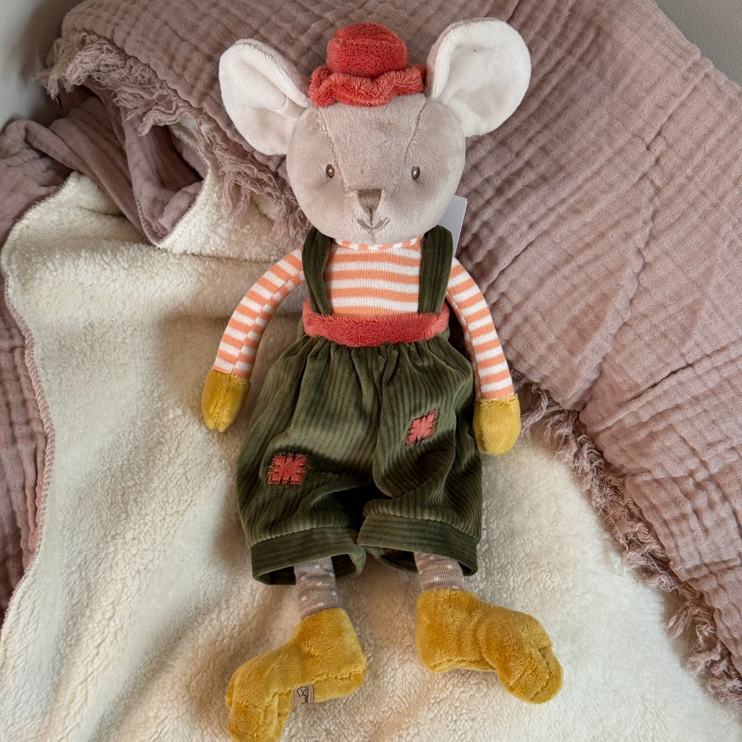 Plush mouse with soft beige fur, floppy ears, and black stitched eyes, wearing a coral velvet hat, coral-and-white striped shirt, green corduroy overalls with a coral patch on each pant leg and a matching coral waistband. The mouse has mustard-colored hands and feet and is lying on a soft white blanket with a mauve fringed textile in the background. 