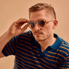 Load image into Gallery viewer, Georgie Toasted Moxie Checkered Sunglasses, person wearing round acetate sunglasses with UVA/UVB protection, gradient amber-brown lenses, and red, cream, brown, and golden yellow checkered frames against beige background. 