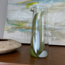 Load image into Gallery viewer, Tall clear glass pitcher with pistachio green and white elongated accents and pour spout, displayed on wooden surface in front of abstract painting. 