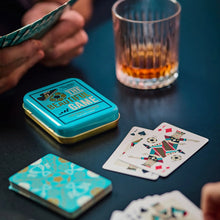 Load image into Gallery viewer, Football Themed Playing Cards, turquoise and gold playing card tin with "THE BEAUTIFUL GAME" lettering displayed beside illustrated playing cards and a ribbed whiskey glass on a dark table during a card game. 