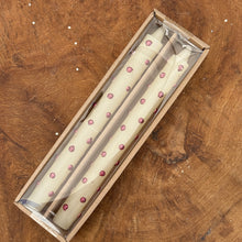 Load image into Gallery viewer, Two white taper candles with red metallic polka dots in a brown packaging box. Box has a clear plastic top. On a brown wooden surface. 