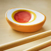 Load image into Gallery viewer, Glossy dolomite chopstick rest shaped like a halved boiled egg with orange and yellow center and mustard yellow exterior, displayed with wooden chopsticks on a neutral background. 